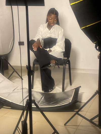A young Black woman with dark hair pulled back is seated on a black chair in a studio setting. She is wearing a white collared shirt and wide-legged black pants, with black and white striped shoes. Her left leg is crossed over her right. She is looking down with a neutral expression.
The studio is equipped with professional lighting equipment. There are large softboxes on stands to the left and right of the subject, and a floor reflector in front of her. The background is a plain white wall, and the floor appears to be tiled. The scene is well-lit, suggesting a professional photoshoot.
The image does not contain any identifiable landmarks or clues that would place the location in South Ropp, Nigeria, or any specific city. The provided information is solely based on the visual content of the image.