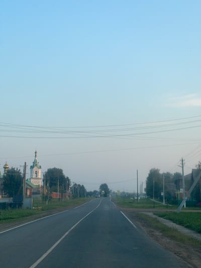 The image shows a road leading through a village. On the left side of the road, there is a church with a green roof and white walls. There are also several houses and trees. On the right side of the road, there are more houses and trees, as well as power lines. The sky is clear and blue. The road is paved and has a white line down the center. There is a car driving on the road in the distance. The scene appears to be peaceful and rural.