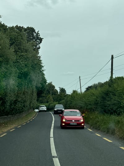 The image shows a view from a vehicle driving on Eyrefield Road in Ireland. The road is asphalt and has a white dashed line and a solid white line separating lanes. The road is bordered by thick green hedges and trees on the left and right. In the distance, there are several cars on the road. The sky is overcast and gray. The red Volkswagen Golf in the foreground has the license plate 161-WX-1599. A dark-colored SUV is behind it, and further ahead, a white SUV and another white car are visible. Utility poles with wires are present on the right side of the road.