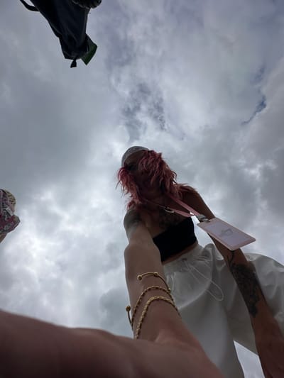 From a low angle shot taken in Kampung Toyapakeh, Indonesia, a woman with pink hair, tattoos, and gold bracelets is looking down while holding a badge. She is wearing a black top and white pants. The sky is cloudy. There's a bag in the lower left and a person in a floral shirt in the lower center.