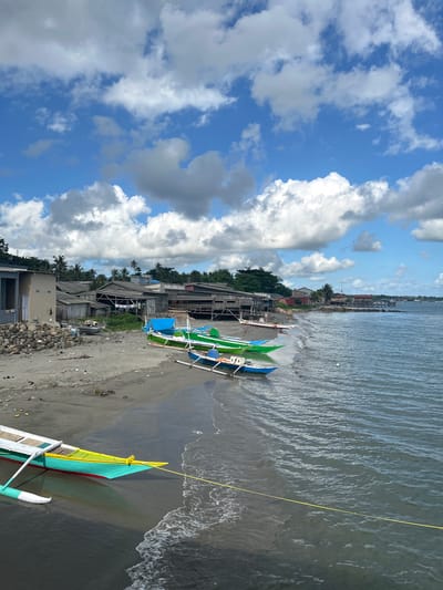 This image depicts a tropical beach scene in Ara, Indonesia. The foreground features several colorful outrigger boats pulled up onto the sandy shore. These boats are predominantly green and blue, with some accented with red and yellow. The water is a calm, dark blue, with gentle waves lapping at the beach. A yellow rope is visible extending from one of the boats into the water.
Behind the boats, a small coastal village lines the shore. The buildings appear to be constructed from wood and tin, with some showing signs of age and weathering. Palm trees are scattered throughout the village, adding to the tropical ambiance. In the background, the horizon is a mix of green vegetation and distant structures.
The sky is a vibrant blue, dotted with large, fluffy white clouds that cast shadows on the landscape. The lighting suggests it is daytime, likely mid-morning or afternoon. The overall mood is peaceful and serene, capturing the essence of a quiet seaside community. No visible text is present in the image.