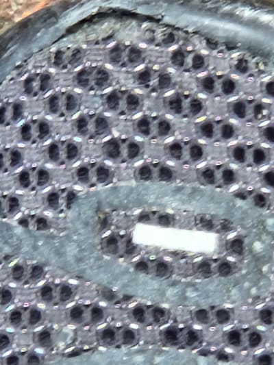 The image is a close-up, macro shot of a black, textured material, likely a fabric or rubber, with a honeycomb or mesh pattern. The material appears to be part of an object, possibly footwear or an accessory, given the visible wear and tear on the left side, where a gray, possibly leather or plastic edge is peeling away.
Within the mesh, there are numerous small, dark circular openings. Some of these openings have bright, white or iridescent highlights, suggesting a reflective quality of the material or a play of light on its surface. In the center of the frame, a rectangular, white element is embedded within the mesh. This element is smooth and contrasts sharply with the textured background. Its purpose is unclear from the image alone, but it could be a logo, a functional component, or a decorative feature.
The overall impression is one of detailed texture and intricate pattern. The lighting is somewhat diffused, creating soft shadows and highlights that emphasize the three-dimensionality of the material. There is no indication of people, animals, or a specific scene; the focus is solely on the material and its texture. The setting is not discernible. No text is visible in the image.
