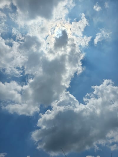 The image depicts a dramatic sky filled with a mix of bright white and darker gray clouds against a vibrant blue sky. Sunlight streams through a gap in the clouds, creating a luminous effect and visible sunbeams. In the lower portion of the image, two construction cranes are visible, suggesting an urban or industrial environment. The perspective is looking upwards. Based on the presence of construction cranes and the general visual aesthetic, this scene is consistent with a view from a developed area within Daejeon, South Korea, likely during daylight hours. There are no people or animals visible in the frame.