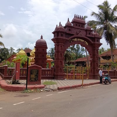 The image depicts a religious site in Marna, India, featuring an ornate, reddish-brown entrance archway inscribed in a non-English script. Adjacent to the archway is a similar-colored wall and architectural detail, including a domed tower. Behind the gate of the temple golden roofs can be seen. A black informational plaque is mounted near the entrance. A man stands next to a parked teal scooter on the side of the road outside the temple. The road is asphalt with faded white lane markings. Palm trees are visible in the background against a partly cloudy sky. The overall scene appears to be a street view of a temple complex.