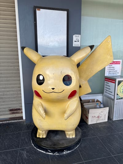 The image shows a large, slightly weathered, yellow statue of Pikachu standing on a black base. The statue is located next to a building with a gray wall, a closed metal security gate, and a white sign with a black frame. Above the Pikachu statue and behind the building is a metal structure with advertisements or signage in Thai, as well as a no-smoking sign. Also visible is a cardboard box with several water bottles. This image was taken in the city of Ban Ko Pru, Thailand.
