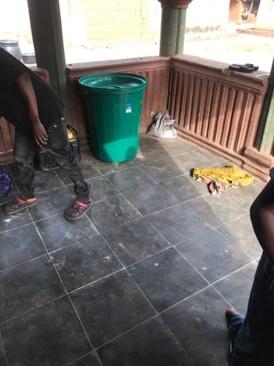 The scene is an outdoor or semi-enclosed tiled area, likely a veranda or patio, located in Ogodo, Nigeria. The dark square tiled floor shows some scuff marks and light residue. Two individuals are partially visible. On the left, a person wears a black short-sleeved shirt, dark jeans, and black clogs with prominent red/orange straps, leaning forward. Behind them, a small yellow generator is situated. A large, ribbed green plastic trash can with a lid stands against a brown wall section. To the right of the trash can, a white sack or bag containing undisclosed contents rests against the base of the brown, vertically ridged wall. A pile of multi-colored fabric, predominantly yellow, lies on the tiled floor in the mid-ground right. On a ledge above the ridged wall, two black sandals are placed. On the far right, the lower leg and foot of a second person, wearing blue jeans and a brown shoe, are visible. The area is under direct sunlight, indicated by strong shadows. In the background, beyond the immediate structure, several large dark pots or containers are discernible.