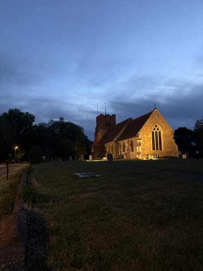 The image depicts a brick church illuminated by warm lighting against a dusky blue sky. The church has a prominent tower and a steep, tiled roof. Several gravestones are visible on the well-maintained lawn in front of the church. To the left, a street is lined with trees and a lamppost. The scene appears to be a graveyard or church grounds. The time of day is dusk or early evening. The location is identified as the city of Chelmsford, United Kingdom.