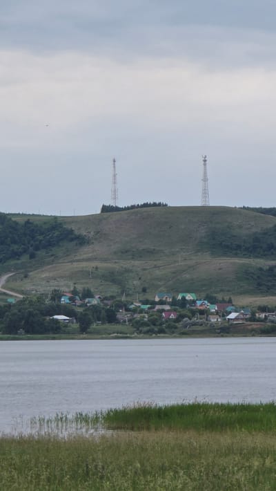 This is a landscape photograph taken from across a body of water, likely a lake or river. In the foreground, tall green and brown grasses are visible, with reeds growing from the water's edge. The water itself is calm and reflects the overcast sky.
In the midground, across the water, a small village is nestled at the base of a rolling green hill. The houses are varied in color and style, with many featuring brightly colored roofs. Trees are interspersed throughout the village and on the lower slopes of the hill. A winding dirt road is visible on the side of the hill, leading up towards the crest.
Dominating the background are two tall, lattice-style communication towers positioned on the summit of the hill. The sky above is heavily overcast with a pale, diffused light. The overall scene suggests a rural or semi-rural setting in the town of Kupoyarovo, Russia.