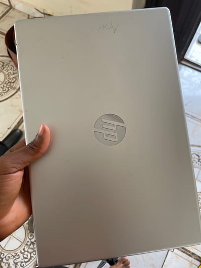 A hand is holding the lid of a silver HP laptop. The HP logo is visible in the center of the lid. The laptop appears to be used, with some minor scuffs and scratches on the surface. The background shows a tiled floor, suggesting an indoor setting, possibly a home or office. A person's hand, with visible fingernails, is supporting the laptop from underneath. The lighting suggests it is daytime. The overall mood is neutral, focusing on the object itself. There is no visible text in the image, other than the HP logo.