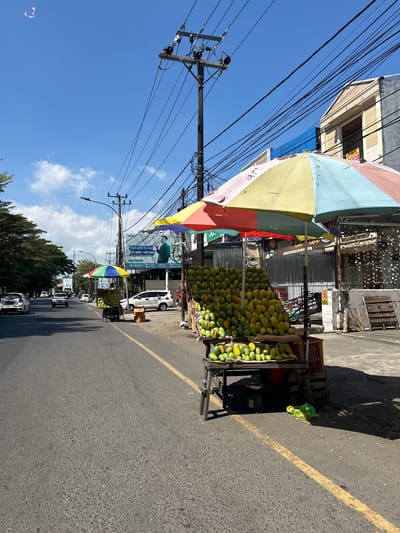 This media file shows a street scene in Makassar, Indonesia, under a bright, sunny sky with some scattered clouds. The image is oriented vertically, so the scene appears sideways.

The primary focus is a roadside fruit stand, prominently featuring a large pile of green and yellow mangoes. The stand is covered by a colorful, multi-panel umbrella, with segments of pink, yellow, light blue, and green. Behind the fruit stand, various buildings and structures line the street, including what appears to be a shop with a corrugated metal facade. Overhead, a dense network of power lines crisscrosses the sky, suspended from utility poles.

In the background, a white car is parked near a billboard advertising "HIDUP LEBIH TENANG BELI ACER-PASTI AMAN" (Live More Peacefully Buy Acer - Definitely Safe) and "Deltasoft." Further down the street, several other vehicles, including cars and what might be motorcycles, are visible, suggesting moderate traffic. The road itself is asphalt with a yellow line marking the lane. Trees are visible along the side of the road in the distance.

The scene appears to be set during the daytime, indicated by the strong sunlight casting shadows and the clear blue sky. The overall impression is of a bustling street market in a tropical urban environment. The visible text "BERAS DIJUAL" (Rice for Sale) suggests other goods might also be available nearby.