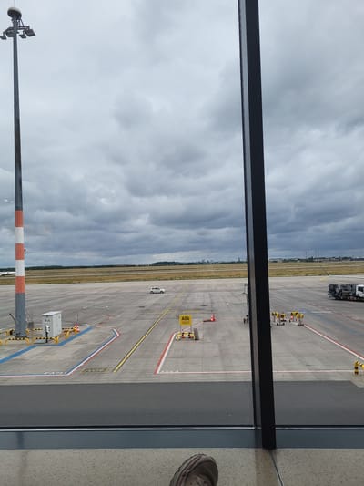 The image shows an airport tarmac under a cloudy sky, as viewed from inside a building through a large window. In the foreground, a portion of a person's shoe and leg are visible at the bottom of the frame. A dark vertical window frame divides the view. On the tarmac, a tall light pole with multiple lamps stands on the left, marked with red and white stripes. To the right of the light pole, there is a white electrical box with warning markings and a yellow barrier. Several cones and yellow safety bollards are scattered across the tarmac. Further into the scene, a white car is driving on the tarmac. Yellow signs with black lettering are visible, including one with "A06" and GPS coordinates. In the distance, there are more light poles and structures, as well as a city skyline under the overcast sky. On the far right of the tarmac, a white truck with equipment is parked. The overall environment appears to be Schönefeld, Germany, given the context of an airport.