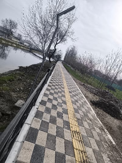 This image displays a long, rectilinear pedestrian pathway in Pakhtakor, Uzbekistan, under an overcast sky. The pathway is constructed from interlocking pavers, forming a checkerboard pattern of alternating light grey and dark grey stones. A yellow tactile paving strip, featuring parallel ridges, runs lengthwise down the center of the path.
To the immediate left of the pathway, a low metal railing or barrier extends into the distance, separating the path from a narrow strip of disturbed earth and sparse vegetation that slopes down to a calm body of water, likely a canal or river. This water reflects the bare deciduous trees lining its opposite bank and the cloudy sky. A black, modern street lamp with an angled arm and a rectangular LED fixture is prominently positioned on the left side, slightly overhanging the pathway.
To the right of the pathway, the ground consists of dirt and scattered, sparse green vegetation. Further right, a substantial pile of dark earth or rubble is visible. A line of bare deciduous trees extends into the distance on this side, with hints of a green fence or netting beyond them. No individuals or active subjects are present in the scene. The leafless state of the trees indicates the season is late autumn, winter, or early spring.