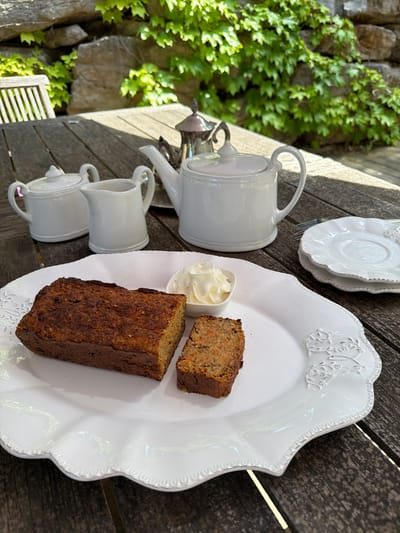 A photograph depicts a still life arrangement, seemingly outdoors on a wooden table, and the image is rotated clockwise roughly 90 degrees. To the left of the frame, there is a stone wall partially covered with ivy. Arranged on the table are two stacked white saucers, a white teapot, two stacked white mugs, and a white decorative plate. On the plate, there are two slices of loaf cake, one smaller than the other, and a small bowl of cream. The photograph appears to be taken in Grimaud, France. The overall impression is that of a tea setting.