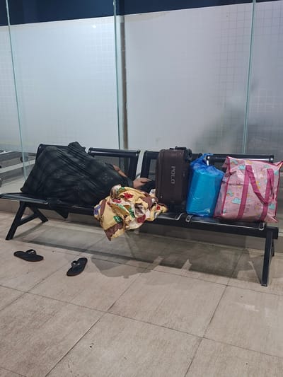 A person is sleeping on a black bench, covered by a dark green blanket with a pattern. Next to the sleeping person, on the bench, are several items: a brown suitcase with "POLO" written on it, a bright blue plastic bag, and a pink tote bag with cartoon cloud and cat designs. A colorful, patterned blanket is also placed on the bench near the person. On the tiled floor in front of the bench are two black flip-flops. The background features a wall with frosted glass panels that have a grid pattern. The scene appears to be an indoor waiting area, possibly at an airport or bus station, given the presence of luggage. The location is specified as Lampeuneurutblang, Indonesia.