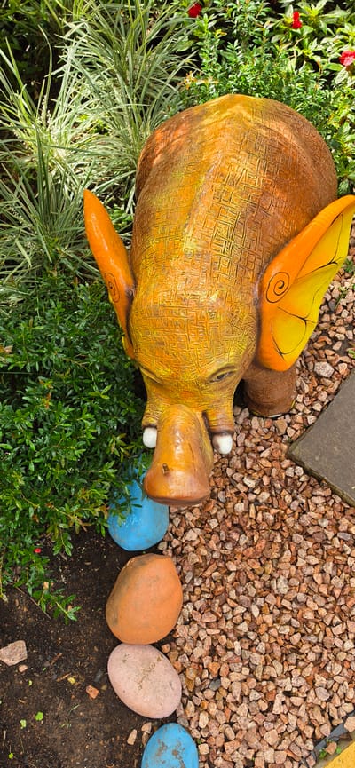 A high-angle shot captures a whimsical, textured duck statue resting among lush greenery, gravel, and painted stones in Gramado, Brazil. The statue is predominantly yellow and brown with orange accents on its beak and one visible wing adorned with a stylized swirl. The surface of the duck is intricately patterned, suggesting a hand-crafted or molded design.
The duck rests amid vibrant green foliage, including grass-like plants with white-edged leaves and small-leafed shrubs. A variety of small, reddish flowers are visible in the background. The surrounding gravel consists of small, reddish-brown stones, next to which are a few larger, painted rocks in shades of blue, pink, and orange, partially buried in the soil. A small, square gray paver sits among the gravel. The scene is lit with natural sunlight, enhancing the texture and colors of the statue and its surroundings.