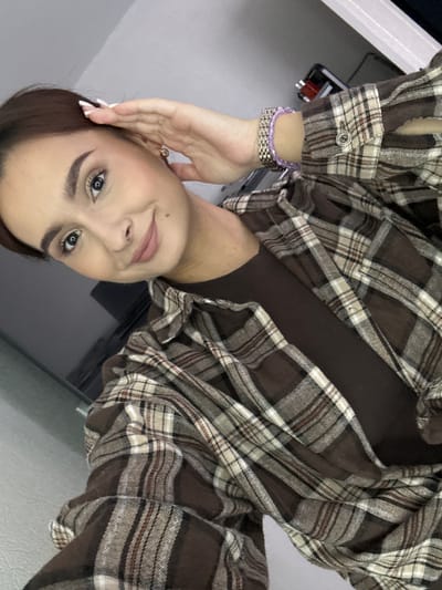 A young woman is captured in a selfie, her face is turned slightly to the right. She has her left hand raised, with her fingers touching her forehead, and she is wearing makeup, including mascara and lipstick. Her hair is dark brown and pulled back. She is wearing a brown t-shirt under an open, brown and beige plaid flannel shirt. On her left wrist, she wears a bracelet with silver and purple beads. The background is out of focus, suggesting an indoor setting, potentially a room or apartment. There is no indication of the location being Kuyuki, Russia.