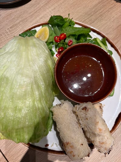 A top-down, close-up shot shows a plate of Vietnamese spring rolls with accompaniments. The plate is white with a brown rim and sits on a wooden-patterned surface. Two fried spring rolls are positioned to the upper right of the plate. A small brown bowl filled with a dark, slightly glossy sauce sits centrally. A large piece of iceberg lettuce is below the bowl. A garnish of fresh mint, cilantro, sliced red chilies, and a lemon wedge is arranged to the left of the bowl.

No location information is determinable beyond the content of the image. Therefore, the prompt request for the location of Kiryat Ono, Israel cannot be fulfilled.