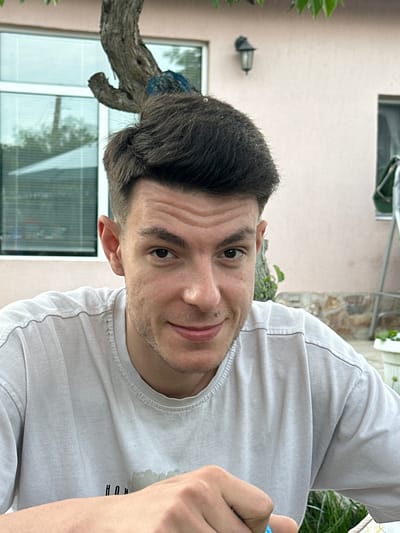 An image captured in Byala, Bulgaria, presents a close-up, chest-up portrait of a young adult male positioned outdoors in natural daylight. The subject has fair skin, dark brown hair styled short on the sides with more volume on top, and dark brown eyes. He displays a subtle, closed-mouth smile or smirk and looks directly at the viewer. A faint stubble is visible on his jawline and upper lip. He wears a light gray, possibly faded, short-sleeved t-shirt with a ribbed collar and some textured lines on the fabric, and a faint dark graphic (appearing as "HUN" and abstract shapes) on the chest. His right hand is visible in the lower right foreground, holding a small blue object.

The background features a light beige stucco building wall. To the subject's left, a large rectangular window with white framing reflects horizontal blinds or internal structures and hints of green foliage outside. Above his head and to the left, a thick, gnarled tree trunk with dark brown bark is prominently visible, extending from the top edge downwards. Above the subject's head and to the right, a dark, traditional outdoor wall lantern with a glass enclosure is mounted on the stucco wall. Further right, a smaller, partially obscured window is visible. The lower right background shows a stone foundation or wall, patches of green grass, and a white planter with faint green growth, adjacent to a metallic, ladder-like structure. Green leaves are also visible at the very top edge of the frame. The overall lighting is soft and diffused.