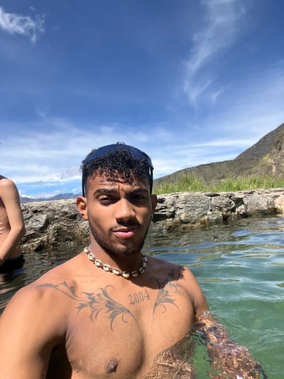 A medium-shot, front-facing selfie shows a young man with dark, curly hair and a beard, wearing sunglasses pushed back on his head and a seashell necklace. He is submerged in clear, green water, with a rocky wall and vegetation in the background, under a bright blue sky with scattered clouds. A portion of another person's torso is visible to the left. The man has tattoos on his chest and left shoulder. The scene appears to be an outdoor natural setting, possibly a hot spring or a natural pool, consistent with the geography of Mucurubá, Venezuela, which is known for its mountainous terrain and natural springs.