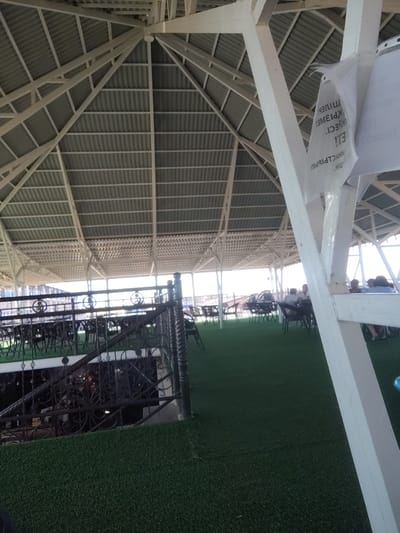 The image depicts an outdoor seating area with a large, white, geometric roof structure supported by columns. The roof is corrugated metal, and the supporting beams form a triangular pattern. White paper sheets with Cyrillic text are attached to a beam in the foreground. The area is covered with artificial turf, and there are several tables and chairs set up. In the background, a group of people are seated at tables. A decorative metal railing is visible in the middle ground, with what appears to be a lower level or stairs behind it. The overall scene suggests a restaurant or cafe with an open-air dining space. The image is captured from a slightly elevated perspective, looking across the seating area towards the background. The location is identified as Toranghylyq, Kazakhstan.