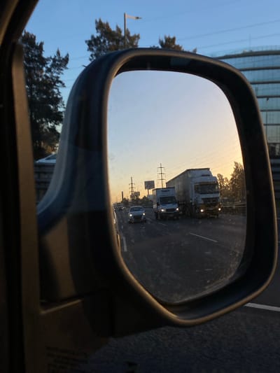 The image shows a car's side mirror reflecting traffic on a highway. The mirror shows a semi-truck, a box truck, and at least one car. Power lines and trees are visible in the background above the highway. The scene appears to be at dusk or dawn. The location is unspecified.