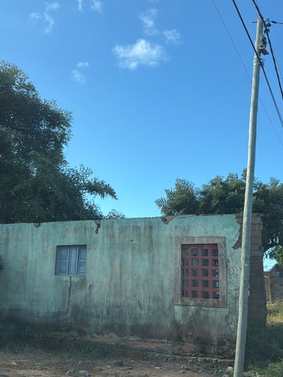 The image depicts a decaying building in Pemba, Mozambique. The building is a light green color with significant weathering and deterioration visible on its exterior. There are two windows on the visible side of the building. The upper window is smaller with red panes, while the lower window is larger with light blue panes. A utility pole and electrical wires are visible in the upper left corner of the image. Trees surround the building on the left side. The ground around the building is barren and covered in dirt. The sky is blue with scattered white clouds.