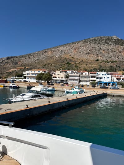 A bright, sunny day is depicted in Gefyra, Greece, with a clear blue sky. In the foreground, a section of a white boat's hull is visible, suggesting the viewer is on board a vessel. The midground features a concrete pier jutting into turquoise water. Several boats are docked along the pier, including a prominent white yacht with a tan canopy. Other smaller boats, some with green hulls, are also present. Behind the pier, a street runs parallel to the water, lined with white buildings that have red-tiled roofs. Cars are parked along the street. A steep, rocky hill covered in dry vegetation rises behind the buildings, dominating the background. The scene conveys a peaceful coastal town atmosphere.
