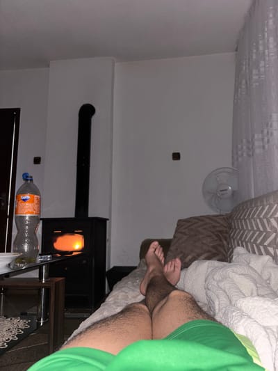 The image shows an interior space. The wall is painted white and features an electrical outlet, a small fan, and a wood-burning stove with a visible chimney pipe. Next to the stove is a side table, upon which sits a partially empty bottle of Fanta soda and other items obscured by the table's edge. In the foreground, a person is lying in a bed with a white comforter and a patterned headboard. The person is wearing green athletic shorts and has hairy legs and feet visible. The location is Grohotno, Bulgaria.