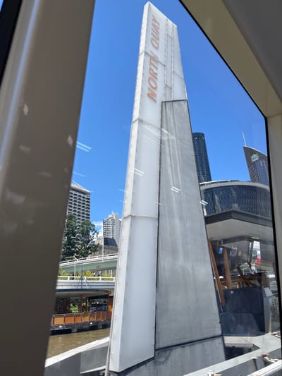 The image presents a bright, clear daytime view of an urban landscape, captured from an interior vantage point looking out through a window. The shot is rotated approximately 90 degrees counter-clockwise from a natural upright perspective.

A prominent white, elongated structure, appearing to be a large sign or part of a modern building, stretches across the central part of the frame. On its visible surface, the text "NORTH QUAY" is displayed in a light orange-brown color.

In the background, a vast, unblemished blue sky occupies much of the upper portion. Several contemporary skyscrapers, featuring glass facades and distinct architectural designs, rise against the sky. Reflections of interior elements or light streaks are visible on the window glass in the foreground.

Below the main structure, the cityscape continues with more buildings, interspersed with green trees, and a body of water, likely a river or canal, visible further down. The overall scene suggests an observation point overlooking a bustling urban area.

No individuals are visible. The clear sky and bright light indicate it is daytime with excellent weather. The text "NORTH QUAY" strongly pinpoints the location to North Quay in Brisbane City, Australia, aligning with the presence of modern city architecture and a prominent waterway.