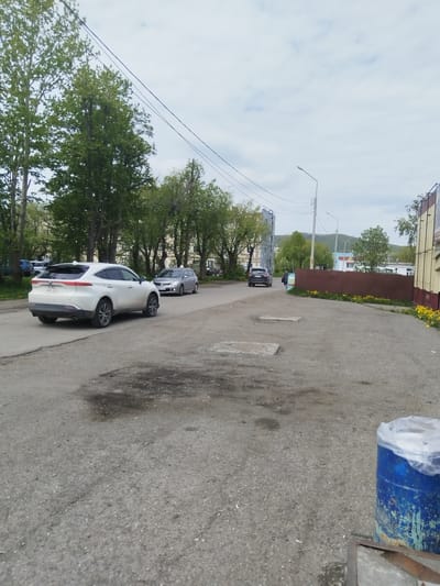 A wide shot shows a road in Vilyuchinsk, Russia, with a white SUV in the foreground. The SUV is parked on the right side of the road, facing left. It is a clean, modern vehicle with tinted windows. In the background, a few other cars are visible, as well as trees and buildings. The sky is overcast. The road is paved, with some potholes and cracks. There is a blue barrel on the right side of the image, covered with a plastic sheet. The overall impression is of a typical urban street scene.