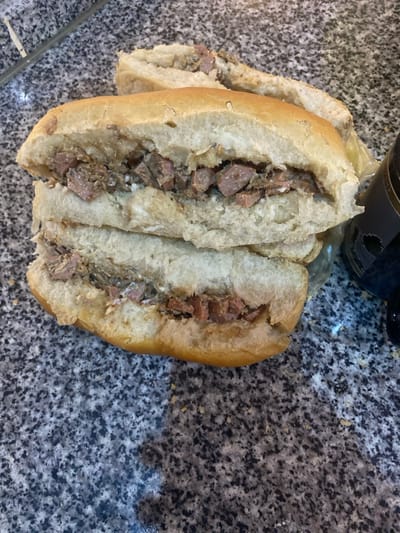 The image displays two open-faced sandwiches or filled bread rolls resting on a speckled grey and black countertop. The rolls are made of light-colored, soft bread, cut lengthwise and generously filled with irregularly diced, cooked meat. The meat appears to be moist and possibly seasoned, exhibiting varying shades of brown and pinkish-brown, consistent with stewed or fried meat. One complete sandwich is prominently featured in the foreground, with another partially visible behind it on the left.

In the upper right background, a dark, possibly glass, bottle is partially visible, with a small section of an unidentifiable label. The countertop surface has a granite-like pattern. There is no human activity depicted, but the prepared food suggests an imminent meal. The lighting is ambient and even, indicating an indoor setting, likely a kitchen or a casual eatery. No discernible text is visible on any objects within the frame. The scene portrays a ready-to-eat meal from Lakka, Sierra Leone, potentially a local street food or a homemade dish.