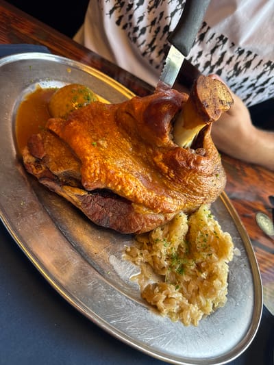 A close-up, high-angle shot shows a large, roasted pork hock served on a silver platter with a side of sauerkraut and a gravy-like sauce. A knife with a black handle is embedded in the pork hock, indicating it is being served or has just been cut into. The pork hock has a crispy, golden-brown skin and a bone protruding from one end. The sauerkraut is light brown and appears to be cooked. The platter has visible scratches, suggesting it is well-used. In the background, a person's arm is visible, wearing a white t-shirt with a black pattern. The scene appears to be set on a wooden table, with a dark blue cloth in the lower left corner. The location is in Fort Lauderdale, United States of America.