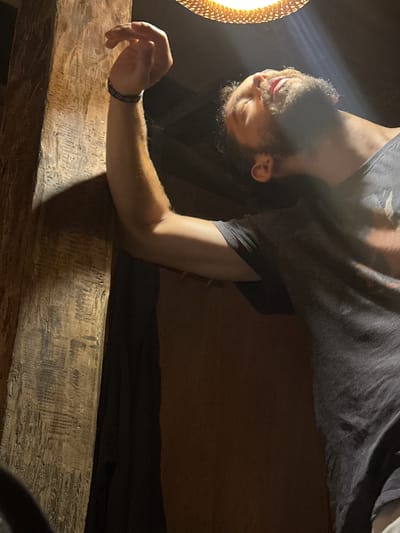 An image, oriented at a 90-degree angle, depicts a man lying down in a dimly lit, indoor setting. The man, with dark, curly hair and a beard, is wearing a dark grey t-shirt. His head is positioned towards the bottom left of the frame, with his eyes closed or mostly closed, and his tongue slightly visible. A bright, warm light source, possibly a lamp with a textured, circular shade, illuminates his face from the left side, casting a glow.

His right arm, bent at the elbow, extends from the upper right and rests upon a thick, rustic wooden beam that spans horizontally across the lower portion of the image. A dark bracelet is visible on his right wrist. The wooden beam exhibits a textured surface, suggesting age or craftsmanship. The background behind the man's body is dark, with subtle hints of a lighter, possibly fabric, surface. The overall atmosphere is one of nighttime or an interior space relying on artificial illumination. The man appears to be in a state of rest or sleep. There is no visible text in the image.