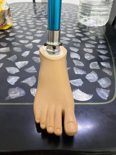 A prosthetic foot is displayed on a dark, patterned surface, likely a table. The foot, made of a light tan material, is connected to a blue metal rod by a silver joint mechanism. In the background, a clear plastic water bottle is visible. A packet of biscuits is located near the bottom left corner of the frame. The setting appears to be indoors. The location is presumed to be Ipoh, Malaysia.