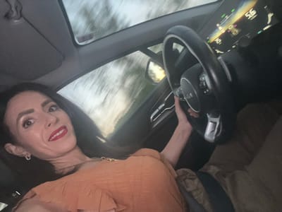 A woman with dark hair and red lipstick is taking a selfie while driving a Kia vehicle. The dashboard display indicates a speed of 95 kilometers per hour. The location is possibly in motion but could not be confirmed as Berg, Luxembourg from the information provided in the images.