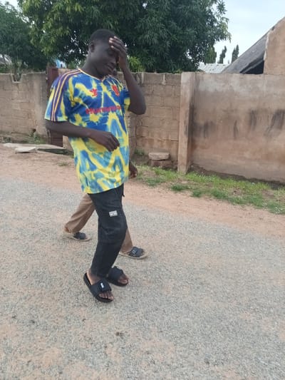 The image depicts an outdoor scene in Zinna, Nigeria. In the foreground, a young Black man is walking and holding his hand to his forehead, suggesting discomfort or heat. He is wearing a blue and yellow tie-dye t-shirt with red accents and the Adidas logo, along with dark jeans and black slide sandals. Behind him, another person, partially visible, is walking in tan pants and light-colored sandals. They are on a gravel road, with a low brick wall and a textured wall on the left and right respectively. Green trees are visible in the background, with the sky overcast. The environment appears to be rural or semi-urban, consistent with a location in Nigeria.