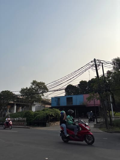 The image shows a street scene in Binong, Indonesia. The perspective is from a low angle, and the image is rotated 90 degrees counter-clockwise, so the top of the image is to the left.

In the foreground, a red scooter carries two people, both wearing helmets. The person in front is wearing a green jacket, and the person behind is wearing a dark shirt. The scooter is on an asphalt road, which takes up the right side of the image. A white line marks the lane. Further down the road, another red scooter is visible, with a single rider wearing a white shirt.

To the left of the road, there are several buildings and vegetation. A two-story building with a pink and blue facade is visible, with a window on the blue section. Overhead, a complex network of electrical wires is suspended from poles, creating a dense web against the pale sky. Trees with green foliage are interspersed throughout the scene, with some branches extending over the buildings and wires. The sky is overcast and bright, suggesting it might be daytime, possibly morning or late afternoon due to the soft lighting.

A sign with white lettering is partially visible on the left side of the road, which reads "TOWN DRIVE" and "MITORY". The overall impression is of a busy, urban or suburban environment.