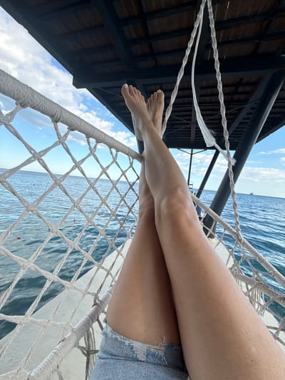 The image shows a person's legs and feet, crossed at the ankles, resting on a net hammock. The hammock is suspended from a wooden structure above, which appears to be part of a pier or boardwalk. The background consists of the calm, blue sea and a cloudy sky. In the distance, a ship is visible on the horizon. The overall scene suggests a relaxed, outdoor setting, possibly a vacation spot. The location is identified as Yalta, Ukraine.