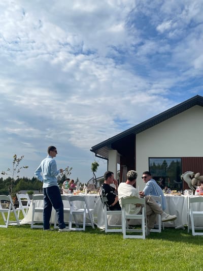 In Simagino, Russia, a group of people are gathered outdoors for a meal at a long table set on a green lawn in front of a modern white house with a dark roof. The sky above is partly cloudy with patches of blue. Several white folding chairs are arranged around the table, which is covered with a white tablecloth and adorned with place settings and some decorative items.

In the foreground, a man in a light blue collared shirt and dark pants stands looking away from the camera. Further back, a group of men are seated at the table, engaged in conversation. One man wears a black shirt, another a cream-colored t-shirt, and a third a light blue and white striped shirt. They appear to be socializing and enjoying the meal. In the background, there are trees and more foliage, suggesting a rural or suburban setting. The overall atmosphere appears to be a casual outdoor gathering.