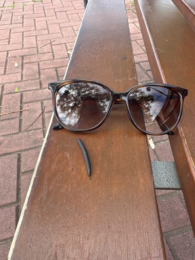 The image depicts a close-up of a brown wooden park bench, partially resting on a brick walkway. A pair of brown-framed sunglasses lies on the bench, reflecting a tree canopy in their lenses. Next to the sunglasses, a millipede crawls on the bench's surface. The location appears to be outdoors but the exact city Esplugues de Llobregat, Spain cannot be confirmed from the image alone. No people are visible.