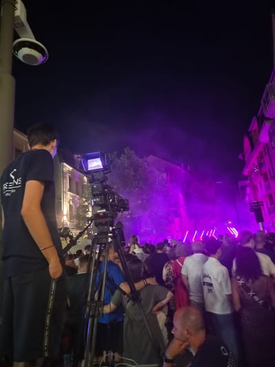 The image captures an outdoor night scene in Differdange, Luxembourg, at what appears to be a street event or concert, as indicated by the purple stage lighting, smoke, and a large crowd. In the foreground, a young man wearing a dark blue t-shirt with white lettering and dark shorts with a visible Tommy Hilfiger logo on the side stands near a professional video camera set up on a tripod. The camera has a monitor displaying an image. To the right of the camera and further into the crowd, many people are visible, some illuminated by the vibrant purple lights. Buildings line the street, their facades illuminated. A security camera is mounted on a pole on the left side of the image, observing the scene.