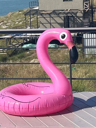 This media file shows an inflatable pink flamingo toy lying on a wooden deck. The flamingo is facing left, with its neck curved upwards and its beak pointing towards the left edge of the frame. It has a black beak and a white eye with a black pupil. The flamingo is partially deflated, with visible creases and folds in the plastic. In the background, there is a black metal fence, behind which is a grassy slope leading down to a body of water. Further in the background, there is a building under construction with scaffolding. The scene is illuminated by bright sunlight, casting a shadow of the flamingo on the deck. The overall impression is of a sunny day, possibly in a coastal or lakeside area. The location is identified as Mysivske rural settlement, Ukraine. No people are visible in the image.