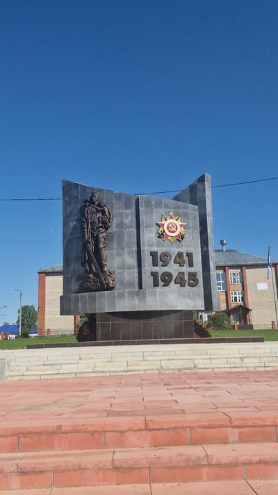 A war memorial in Otchor, Russia is shown. The memorial has the dates 1941 and 1945 displayed vertically, a Soviet star, and a figure of a reclining soldier on it. The sky is visible in the background. A brick walkway is in the foreground. A building is in the background.