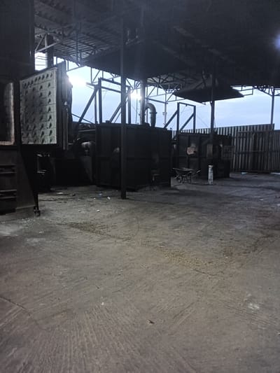 An outdoor industrial scene in the city of Rassvet, Kazakhstan, depicts a large, open-air structure with a corrugated metal roof supported by metal beams and pillars. The ground is a rough, concrete surface with visible cracks and debris. In the foreground, the concrete floor exhibits textured patterns. To the left, a large, dark metal structure is present, possibly a furnace or industrial equipment, with a patterned surface on its side. In the midground, several dark, large, metal containers or structures are visible. A wheelbarrow is positioned to the left of center, in front of one of these containers. In the background, a corrugated metal fence runs horizontally, separating the industrial area from the sky. The sky above is overcast and appears to be either dusk or dawn. The overall impression is one of a utilitarian, possibly disused, industrial workspace.