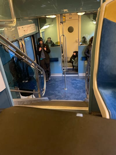 The scene depicts the interior of a bi-level commuter train, consistent with those operating in the Île-de-France region, serving areas such as Saint-Cyr-l'École, France. The vantage point is from an upper deck, looking down two steps into a lower section of the train car. The interior features a consistent blue and off-white color scheme, with blue upholstered seating visible on the right and a dark blue patterned floor on the lower level. Silver metal handrails are present along the stairs and centrally on the lower floor.

On the left, a young adult male with dark hair and a beard stands, angled slightly right. He is dressed in a dark hooded jacket, a lighter shirt with a visible turquoise collar, dark pants, and dark sneakers featuring white soles and a patterned upper. A faint dark reflection, possibly of another person, is visible in a window or reflective panel to his left.

In the background, beyond a cream-colored partition wall that includes a circular window and a closed door, two additional individuals are seated. On the left, a partially visible older male with short grey hair and a beard is seated. On the right, a partially visible female with dark hair is seated, holding a light-colored bag. Both appear to be looking forward.

The floor of the lower section exhibits variations in its dark blue tone, possibly indicating wear or moisture. Artificial overhead lighting illuminates the compartment. A small white sign with red text is affixed to the wall above an open doorway on the left side of the lower level. No direct interactions between individuals are observed.