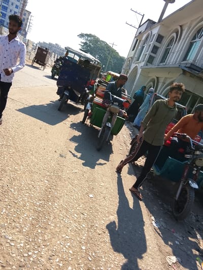 The image displays a street scene in Shibpur, Bangladesh, under bright daylight with strong shadows indicating direct sunlight from a low angle. The foreground consists of a coarse, light-colored asphalt or dirt road surface. On the left, a young male wearing a white, patterned long-sleeved shirt and dark trousers walks towards the left of the frame. In the midground, multiple motorized three-wheeled vehicles, resembling auto-rickshaws or easy-bikes, are positioned along the road. One prominent green vehicle has a male driver in a dark shirt and red cap. Another vehicle behind it is covered with blue fabric. On the right, a young male with a short beard and dark, dishevelled hair, wearing a khaki-green long-sleeved shirt and dark trousers with light side stripes, walks towards the camera, maintaining direct eye contact. A male in an orange shirt is partially visible behind him on another three-wheeled vehicle. In the background on the right, a multi-story building with distinct arched windows and white decorative architectural elements is visible. Utility poles and overhead wires are present near this building. On the far left background, a multi-story building under construction or an incomplete structure is visible, with trees beyond it. Pedestrians, including women in traditional attire, are also visible near the building on the right.