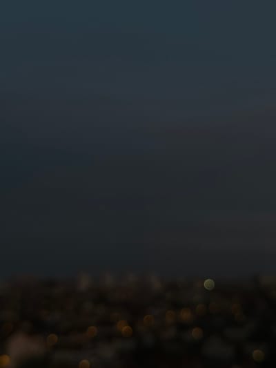 The image depicts a highly blurred, elevated view of an urban area at night or deep dusk in Maisons-Alfort, France. The upper two-thirds of the frame consists of a dark blue-grey sky with no discernible features. The lower third presents a cityscape rendered almost entirely as bokeh, showing numerous indistinct, circular lights against a dark, undefined background. These lights are primarily yellow and orange, consistent with typical urban illumination. A single, more prominent, greenish-yellow circular light is visible slightly above the main cluster of lights in the mid-right portion of the blurred city expanse. No specific structures, individuals, objects, actions, or interactions are distinguishable.