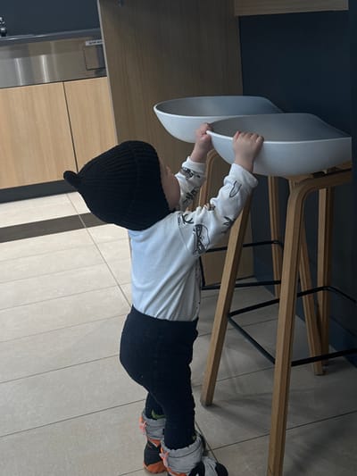 An indoor scene in Ivashevo, Russia, captures a young child standing and facing right, with both hands grasping the front edge of a white, saddle-shaped bar stool seat for support. The child wears a black knitted hat with a pointed top, a long-sleeved white shirt patterned with dark shapes, black textured pants, and grey and orange winter boots. To the child's left and in the background, a light wood counter with a darker surface is visible, above which a stainless steel appliance, possibly a water cooler or coffee machine, is partially seen. The floor is composed of light-colored rectangular tiles. Two identical bar stools, featuring light wooden frames and black metal footrests, are positioned to the right. The immediate background behind the stools consists of a dark, uniform vertical panel or wall. The environment appears to be a modern commercial or public space, such as a cafe or shared lounge area.