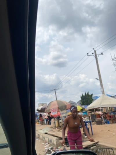 The image depicts a street scene in Naze, Nigeria, captured from a vehicle's interior, indicated by a dark gray structural element framing the left side. The sky is overcast with prominent cumulus clouds.
In the foreground, a young adult female walks from right to left, facing generally forward. She wears a brown short-sleeved shirt, dark red trousers, and a patterned headband. Her arms are bent, and her hands are positioned near an object obscured by the vehicle's lower frame, possibly a wheeled device or small cart. She stands on a dirt ground surface bordering a concrete curb and an open drainage channel containing scattered refuse.
The midground features a commercial strip with multiple market stalls. A prominent stall utilizes a striped orange and off-white/beige parasol with "POS SERVICES" and "POS" text visible. Other stalls feature white canopies. Several individuals are present among these stalls, some seated. Utility poles with multiple electrical lines span the scene, and green foliage is visible behind the market structures. The background reveals additional buildings with varying roof colors and scaffolding on the far right.