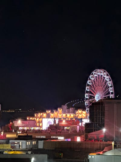 The image shows a night scene at Parque das Nações, Lisbon, Portugal. A large Ferris wheel, illuminated in pink and white, is prominent on the right side of the frame. In the background, a waterfront area is visible with distant city lights. In the mid-ground, there are various structures, including stages or stalls, decorated with bright lights and some appearing to be banners or screens. A crowd of people is gathered in front of these structures. The foreground consists of more structures, some appearing to be temporary buildings or barriers, with lighting elements. The overall scene depicts a vibrant event or festival.